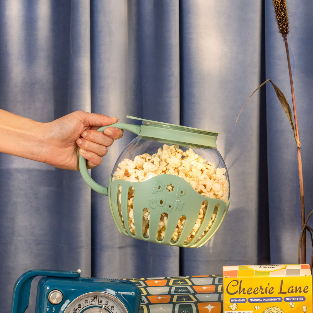 A hand holding a green popcorn popper filled with popcorn in front of a blue velvet curtain