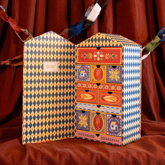 A decorative gift box with an open hinged front, featuring a blue and yellow harlequin pattern on the outer panels and a colorful front drawer section with red, orange, and blue illustrated compartments. The box is displayed on draped brown fabric in front of a matching brown curtain backdrop.