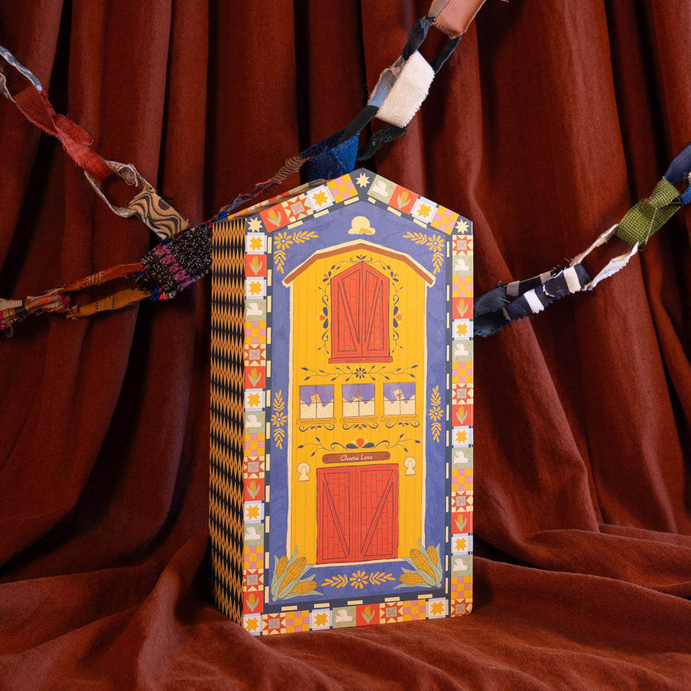 A decorative hand-painted wooden box shaped like a small house or barn, featuring a blue and yellow facade with red doors and floral borders, set against a draped brown fabric background.