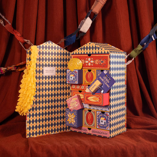 A decorative gift box set displayed against a red curtain backdrop. The set includes a large blue-and-yellow harlequin-patterned box with a yellow ruffled trim, an open matching box showing multiple smaller colorful boxes inside, and a small red box labeled 'Meringue Crème'.