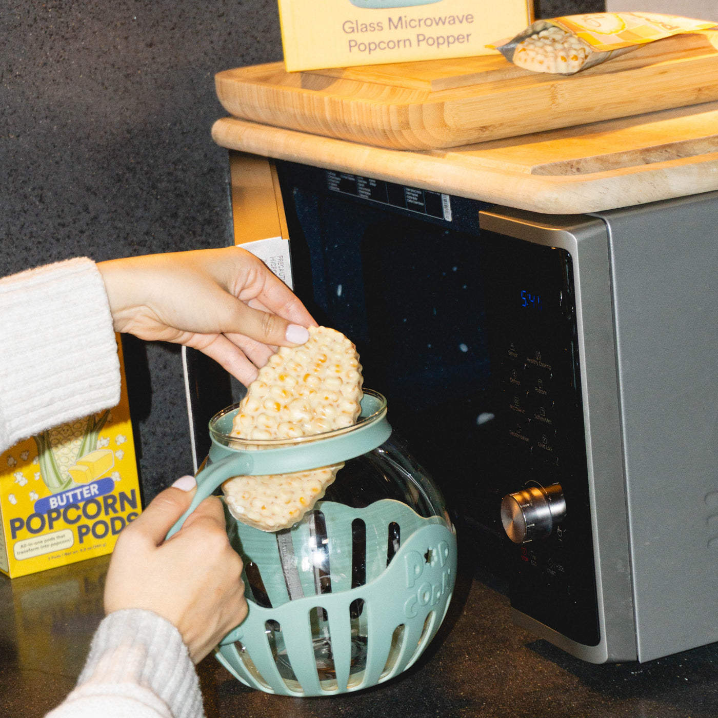 Person using a microwave popcorn popper with a box of popcorn pods in the background.