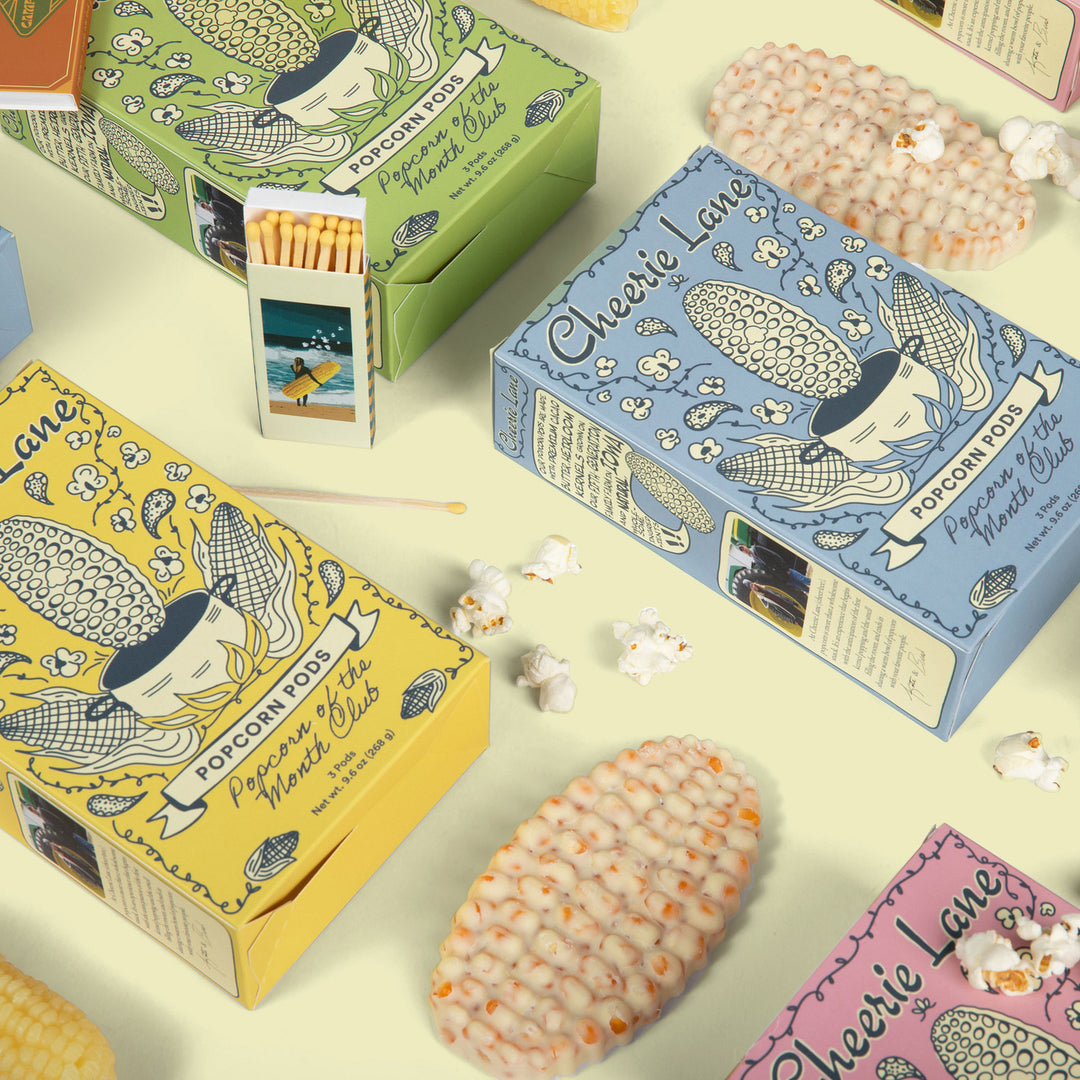 Colorful boxes of Cheerie Lane popcorn with popcorn kernels on a light background