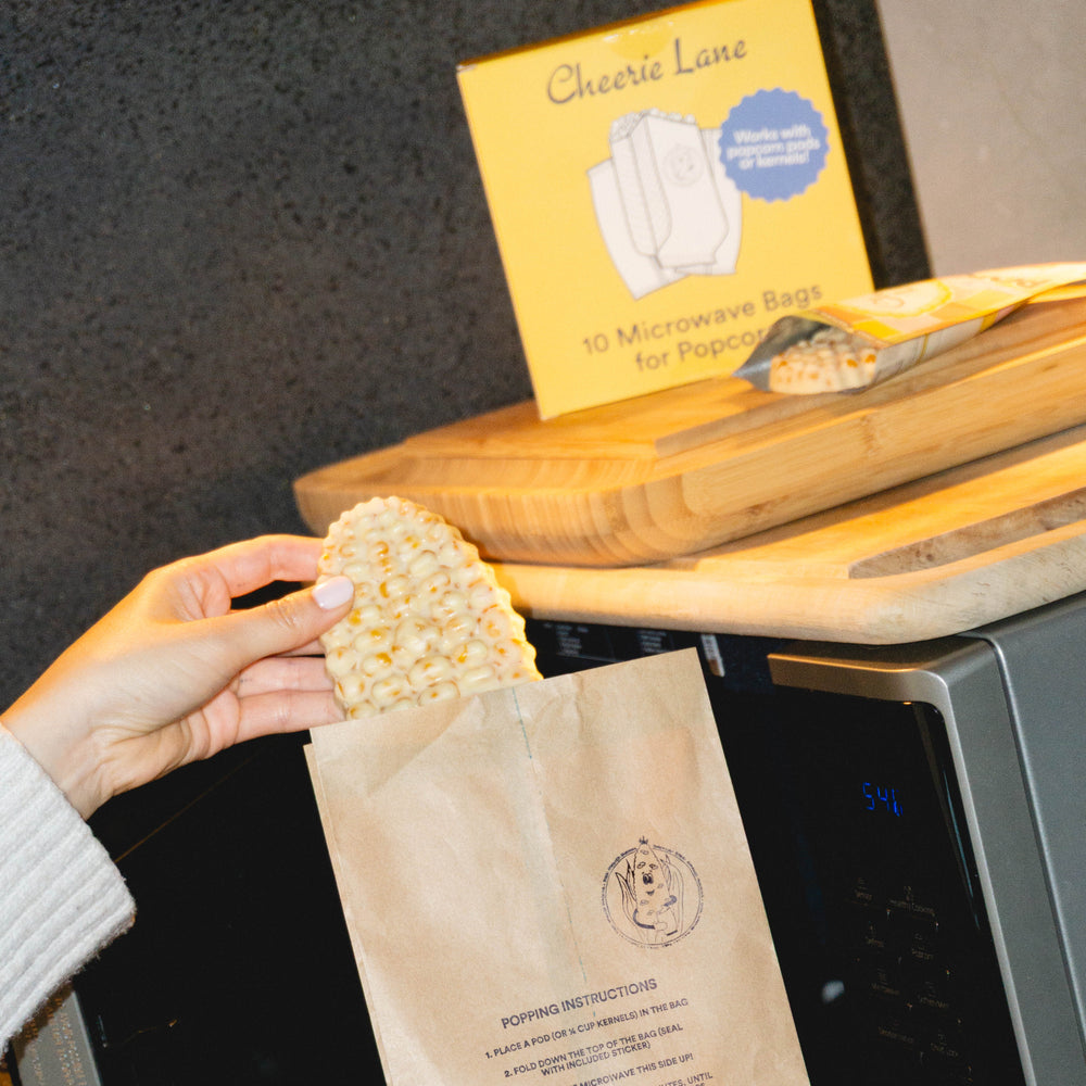 Person holding a brown paper bag with popcorn and a box of Cheerie Lane popcorn bags in front of a microwave.
