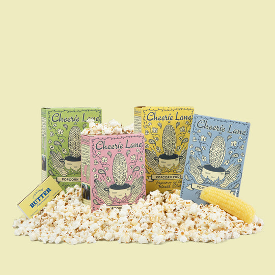 Four colorful popcorn tins from Cheerie Lane with popcorn and a corn kernel on a light yellow background.