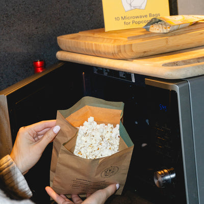 Person holding a bag of popcorn in front of an open microwave with a box of popcorn bags on a wooden surface.